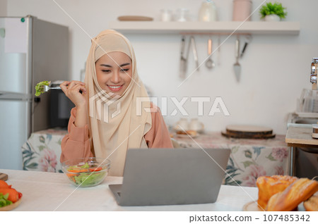 Beautiful Asian Muslim woman is eating her healthy salad while working on her laptop in the kitchen 107485342