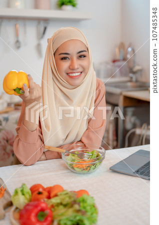 Beautiful Asian Muslim woman in a hijab sits at a kitchen table with a sweet pepper in her hand Beautiful Asian Muslim woman in a hijab sits at a kitchen table with a sweet pepper in her hand 107485348