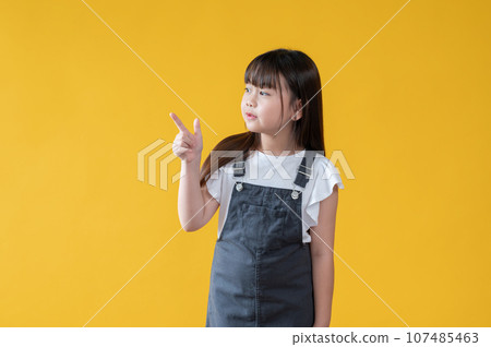 An adorable little Asian girl in a cute jeans dress is looking and pointing her finger aside 107485463