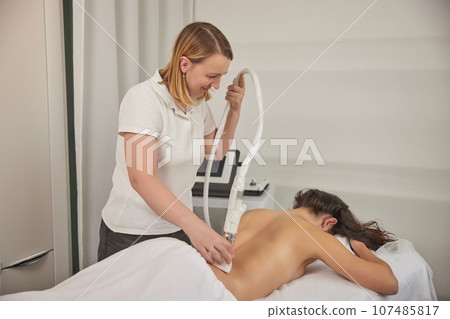vacuum-roller manipulator, beauty clinic, anti cellulite. Attractive young woman is in process of improving body shape, using vacuum roller to massage client, correcting figure, anti-cellulite massage 107485817