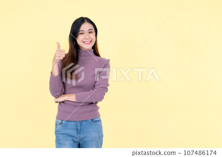 A beautiful Asian woman shows her thumb up and stands against an isolated yellow background 107486174