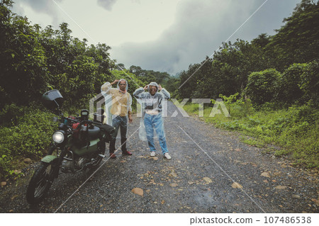 couples of asian biker wearing plastic rain clothes standing beside small enduro motorcycle on country track 107486538