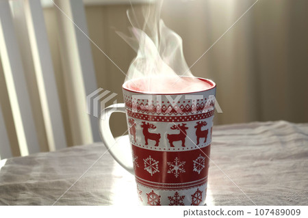 Steaming Cup of Coffee or Tea on Table. White linen Tablecloth. Close up ceramic red Christmas mug with smoke on old table in dining room interior background. Hot Drink Concept. Cozy Morning. 107489009