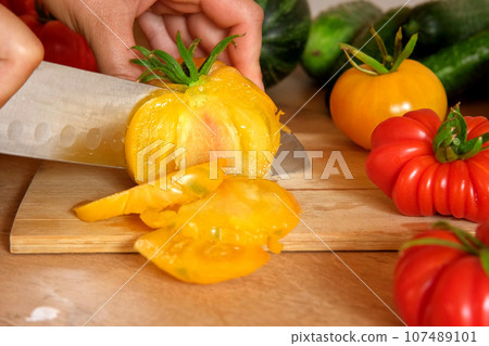 Woman using a knife cutting fresh raw vegetables. Tomatoes, cucumbers being sliced on a wooden board. Cooker at home making a healthy veggie meal. Chopping ingredients for preparing vegetarian dish 107489101
