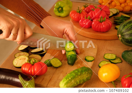 Woman using a knife cutting fresh raw vegetables. Tomatoes, cucumbers being sliced on a wooden board. Cooker at home making a healthy veggie meal. Chopping ingredients for preparing vegetarian dish 107489102