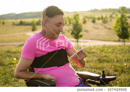 A young gay man in a wheelchair using a mobile phone in the park at sunset. 107489906