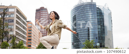 Vertical shot of young asian woman posing happy, raising hands up and dancing, triumphing, celebrating victory, enjoying day out in city 107490612