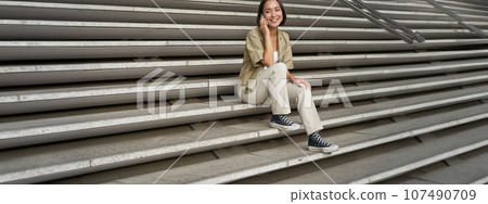Smiling asian girl sits on stairs of building and talks on mobile phone, relaxing during telephone conversation 107490709