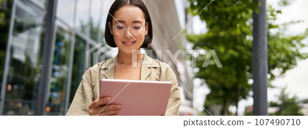Outdoor shot of young korean woman stands on street with laptop, wears glasses, reads, smiles happily 107490710