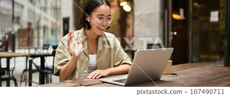 Portrait of young woman video chatting, having online meeting on laptop, sitting in outdoor cafe with compute, wearing glasses and smiling 107490711