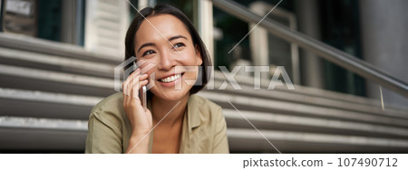 Portrait of asian girl smiles while talks on mobile phone. Young woman calling a friend, sitting on stairs Portrait of asian girl smiles while talks on mobile phone. Young woman calling a friend, sitting on stairs 107490712