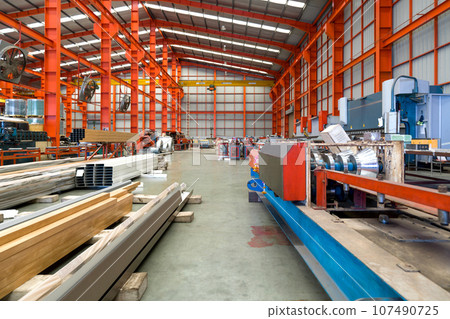 A gargantuan warehouse brims with a labyrinth of gleaming steel beams crisscrossing overhead, creating a metallic canopy. There are steel sheet roll and steel tube on the floor. 107490725