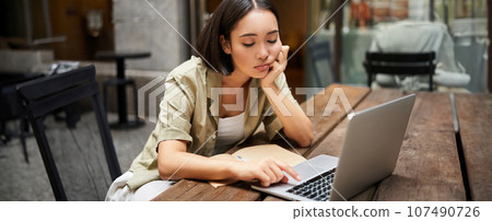 Bored asian girl sitting in cafe and staring at laptop, studying and feeling sad, doing homework 107490726