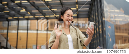 Happy asian girl waves at her smartphone camera, says hello to friend on video chat, calling someone, standing on street Happy asian girl waves at her smartphone camera, says hello to friend on video chat, calling someone, standing on street 107490753