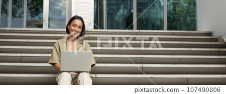 Young asian woman using laptop outdoors. Cute girl student with computer, does her homework, types, sits on stairs outside campus Young asian woman using laptop outdoors. Cute girl student with computer, does her homework, types, sits on stairs outside campus 107490806