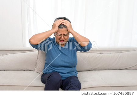 A distressed woman sits on a plush sofa, hands clasped atop her head. Her eyes, full of worry, lost in thought. The quiet room echoes her mood, evoking a sense of solitude. 107490897