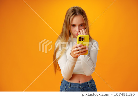 Surprised teen girl using mobile phone against yellow background Surprised teen girl using mobile phone against yellow background 107491180
