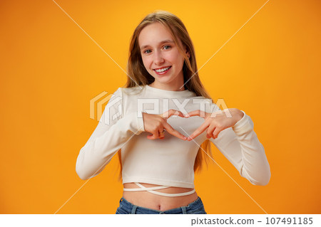 Friendly teen girl shows heart love gesture on yellow background Friendly teen girl shows heart love gesture on yellow background 107491185