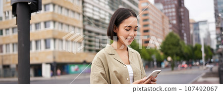 Young asian woman exploring city with smartphone app, holding mobile phone and walking on screet 107492067