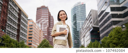 Portrait of young asian woman, looking happy and confident, going to work or university, city skyscrappers behind her, holding laptop and notebook 107492084
