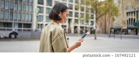 Portrait of stylish urban girl walks in city with tablet, drinks takeaway coffee 107492121