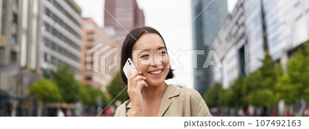 Portrait of smiling asian woman talking on mobile phone, walking on street near busy road and speaking to friend, laughing Portrait of smiling asian woman talking on mobile phone, walking on street near busy road and speaking to friend, laughing 107492163