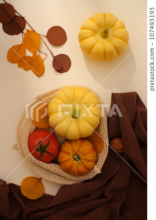 Top view of a mesh basket of just harvested fruit decorated with dark brown fabric and dried autumn leaves on a light beige background. Autumn concept with a bountiful harvest 107493193