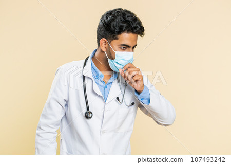 Indian young doctor man in protective mask cough with seasonal flu symptoms fever allergy infection Indian young doctor man in protective mask cough with seasonal flu symptoms fever allergy infection 107493242