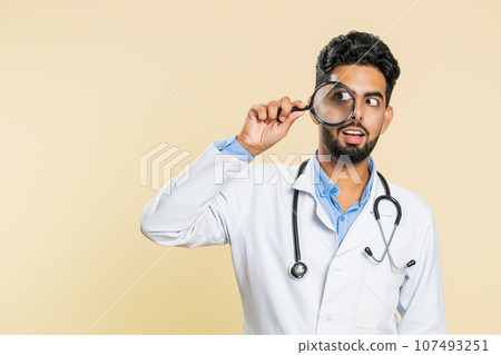 Indian doctor man holds magnifying glass near face looking at camera with big zoomed eye, analyzing 107493251