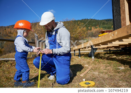 Father with toddler son building wooden frame house on pile foundation. Male builder and kid playing with tape measure on construction site on sunny day. Carpentry and family concept. 107494219