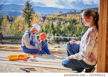 Father, mother and son building wooden frame house. Toddler boy helping his daddy, while woman looking for them on construction site. Guys wearing helmet and blue overalls. Carpentry, family concept. 107494222