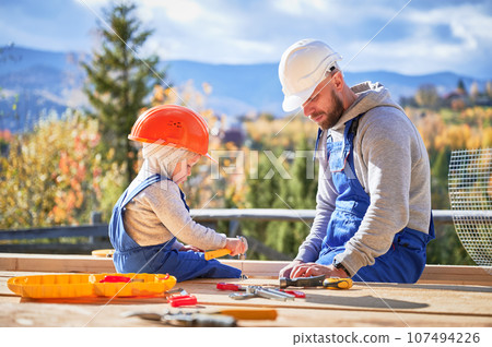 Father with toddler son building wooden frame house. Boy helping his daddy, hammering nail into plank on construction site, wearing helmet and blue overalls on sunny day. Carpentry and family concept. 107494226