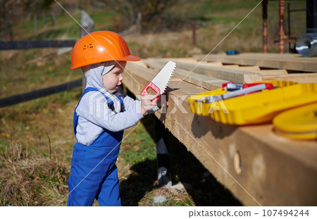 Boy toddler playing as builder on construction site. Child carpenter in orange helmet and blue overalls learning to build wooden frame house outdoor on sunny day. Carpentry and workshop concept. Boy toddler playing as builder on construction site. Child carpenter in orange helmet and blue overalls learning to build wooden frame house outdoor on sunny day. Carpentry and workshop concept. 107494244