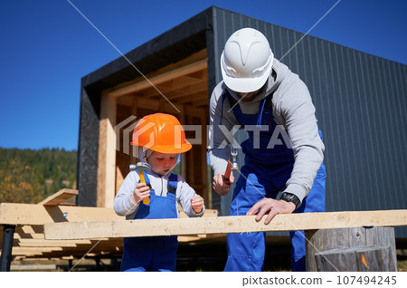 Father with toddler son building wooden frame house. Boy helping his daddy, hammering nail into plank on construction site, wearing helmet and blue overalls on sunny day. Carpentry and family concept. Father with toddler son building wooden frame house. Boy helping his daddy, hammering nail into plank on construction site, wearing helmet and blue overalls on sunny day. Carpentry and family concept. 107494245