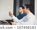 Side view, waist up two caucasian male programmers sitting by the office desk discussing something 107498183