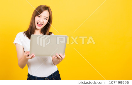 Asian happy portrait beautiful cute young woman teen smile standing wear t-shirt hold laptop computer and excited celebrating success looking to camera, studio shot yellow background with copy space Asian happy portrait beautiful cute young woman teen smile standing wear t-shirt hold laptop computer and excited celebrating success looking to camera, studio shot yellow background with copy space 107499996