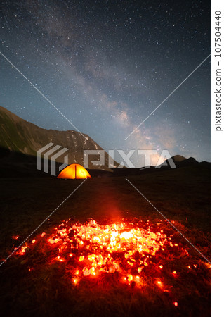 Night landscape long exposure. burning coals are the remains of a fire against the backdrop of a luminous trekking tent of the mountains and the milky way Night landscape long exposure. burning coals are the remains of a fire against the backdrop of a luminous trekking tent of the mountains and the milky way 107504440