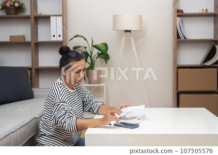 Housewife calculates household finances on smartphone calculator at light table in flat room woman works with documents with utility prices for month stay in apartment 107505576