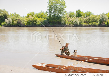 Pensive male fisherman with big rod looking away, while sitting on stern of old boat at pier. Front view of calm man in hat fishing with dog companion, while resting on weekend. Concept of fishery. Pensive male fisherman with big rod looking away, while sitting on stern of old boat at pier. Front view of calm man in hat fishing with dog companion, while resting on weekend. Concept of fishery. 107507015