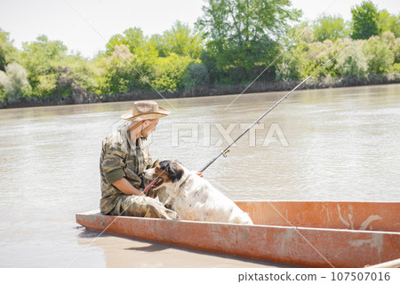 Thoughtful male angler with cute dog holding fishing rod, having rest, while floating on old vessel. Side view of tired man in camouflage fishing, with bushy coast on background. Concept of fishery. 107507016