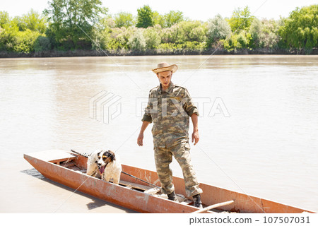Young caucasian guy standing in shubby boat collecting tackle after fishery outdoors. Front view of male angler floating with dog, with calm, muddy river on background. Concept of fishery. Young caucasian guy standing in shubby boat collecting tackle after fishery outdoors. Front view of male angler floating with dog, with calm, muddy river on background. Concept of fishery. 107507031