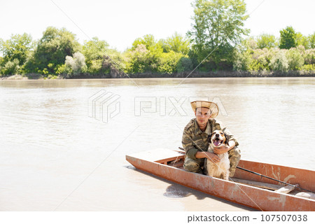 Caring young man in camo posing at camera, while hugging nice spotted setter, while sitting in boat on calm river. Front view of male pet owner petting cute dog, while fishing. Concept of association. 107507038