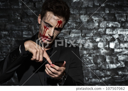 Portrait of a zombie businessman using his smartphone, wearing a black suit, with blood on his face standing against black brick wall 107507062