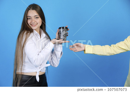 Cheerful pretty girl sincerely looking at camera, while raising paper coffee cup, demonstrating it, with crop anonymous hand beside, isolated on blue studio background. Concept of advertising. Cheerful pretty girl sincerely looking at camera, while raising paper coffee cup, demonstrating it, with crop anonymous hand beside, isolated on blue studio background. Concept of advertising. 107507073
