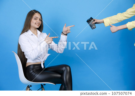 Happy cute lady in leggings pointing at coffee cup, while sitting on chair. Crop view of anonymous hand showing thumb up, while holding paper cup, isolated on blue background. Concept of approval. 107507074