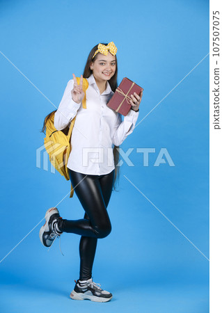 Joyful girl wearing funny headband gesturing, showing victory sign, while smiling at camera. Side view of happy lady with books posing, flirty bending leg on knee, isolated on blue. Concept of posing. 107507075