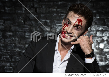 Portrait of a happy white-collar worker holding his hand like a phone near his ear, smiling with zombie bloody make up on his face 107507124