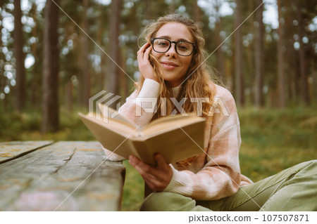 Happy woman is sitting at table with book in beautiful forest. Concept of relaxation, lifestyle. Happy woman is sitting at table with book in beautiful forest. Concept of relaxation, lifestyle. 107507871
