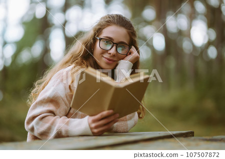 Happy woman is sitting at table with book in beautiful forest. Concept of relaxation, lifestyle. Happy woman is sitting at table with book in beautiful forest. Concept of relaxation, lifestyle. 107507872