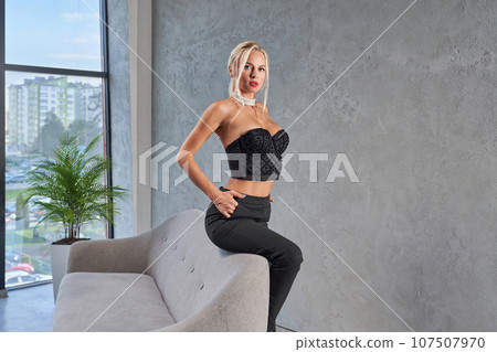 Stunning blonde female in designed strapless top posing, while leaning on sofa edge at home. Side view of pretty woman with hairstyle sitting seductively on couch with hand in pocket. Fashion concept. Stunning blonde female in designed strapless top posing, while leaning on sofa edge at home. Side view of pretty woman with hairstyle sitting seductively on couch with hand in pocket. Fashion concept. 107507970